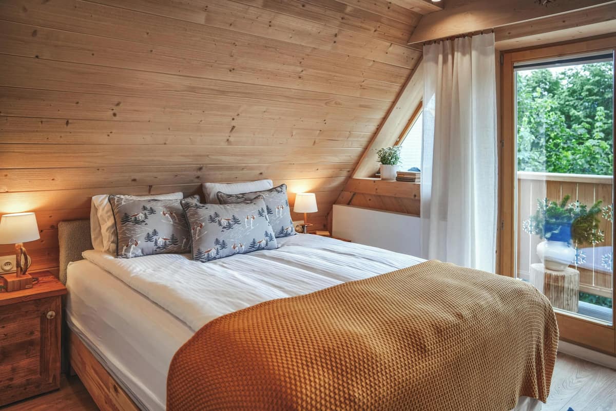 Une chambre confortable avec un plafond en bois, un grand lit agrémenté d’oreillers à motifs, une table de chevet avec une lampe et une fenêtre donnant sur un paysage verdoyant.