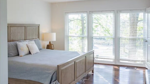 A bedroom with a bed, a nightstand, a lamp, and large windows with blinds.