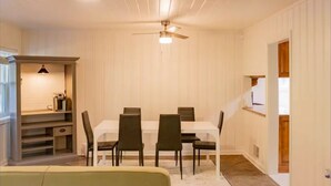 A dining area with a white table and six chairs, a ceiling fan, and a small shelf with a lamp.