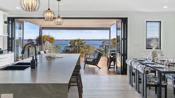 A modern kitchen with a large island, a dining area with a view, and a balcony overlooking the sea. - Modern 4BR Mirraboo Home with Panoramic Lake Views (Mirrabooka)