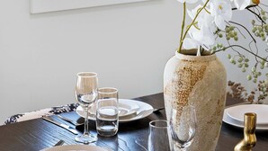 A dining table with plates, glasses, and cutlery, a vase with white flowers, and a window with a view of a body of water. - Modern 4BR Mirraboo Home with Panoramic Lake Views (Mirrabooka)