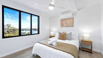 Et soveværelse med et stort vindue, en loftventilator, en seng med hvide sengetøj, to sengeborde med lamper, og en aircondition.