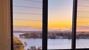 A room with a large window, a chair, a small table with a vase, and a blanket. - Modern 4BR Mirraboo Home with Panoramic Lake Views (Mirrabooka)