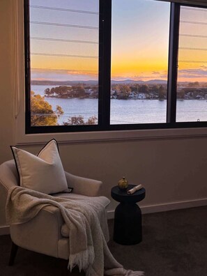 A room with a large window, a chair, a small table with a vase, and a blanket.