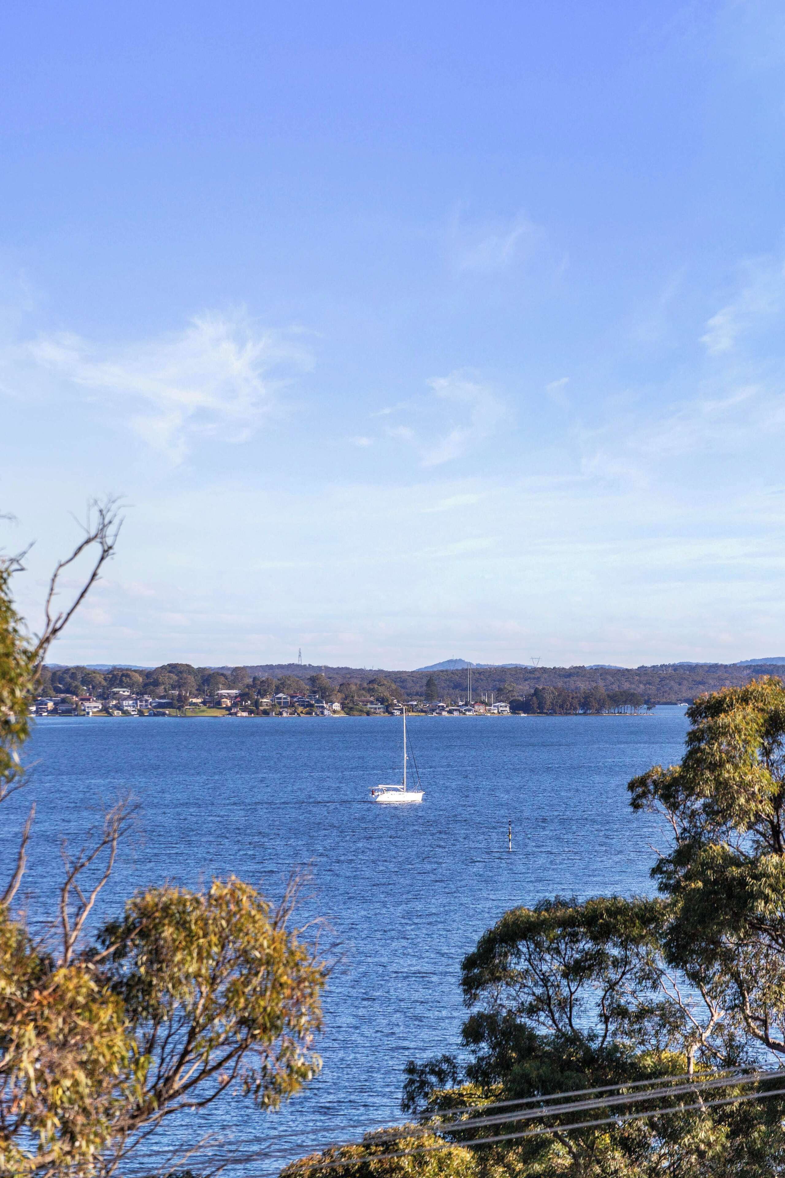 A lake with a sailboat, surrounded by trees and a distant shoreline with buildings.