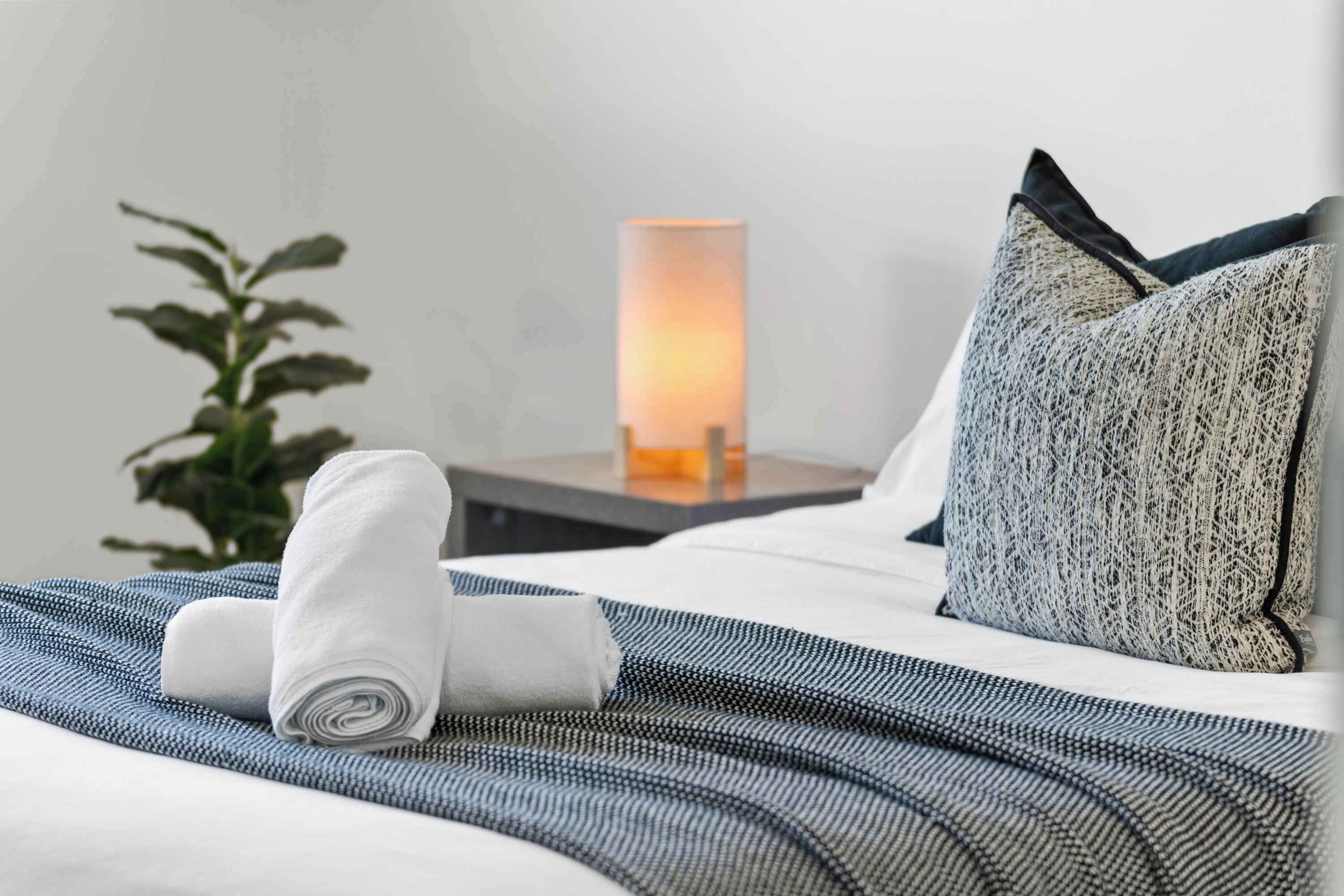 Una cama bien tendida con una toalla blanca enrollada, un cojín texturizado y una lámpara de noche.