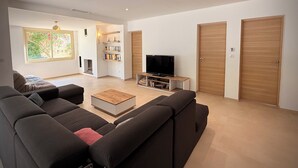 A modern living room with a sofa set, a coffee table, a TV on a wooden stand, and a bookshelf. There is a window with a view of greenery and a door to the right. - Maison Sud: Between sea and vineyards (Martigues)