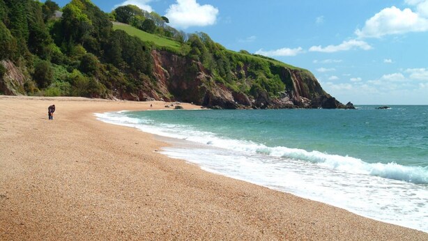 A pebbled beach with a person walking, a cliff with greenery, and the ocean. - The Most Beautiful and Cosy House (Kingsbridge)