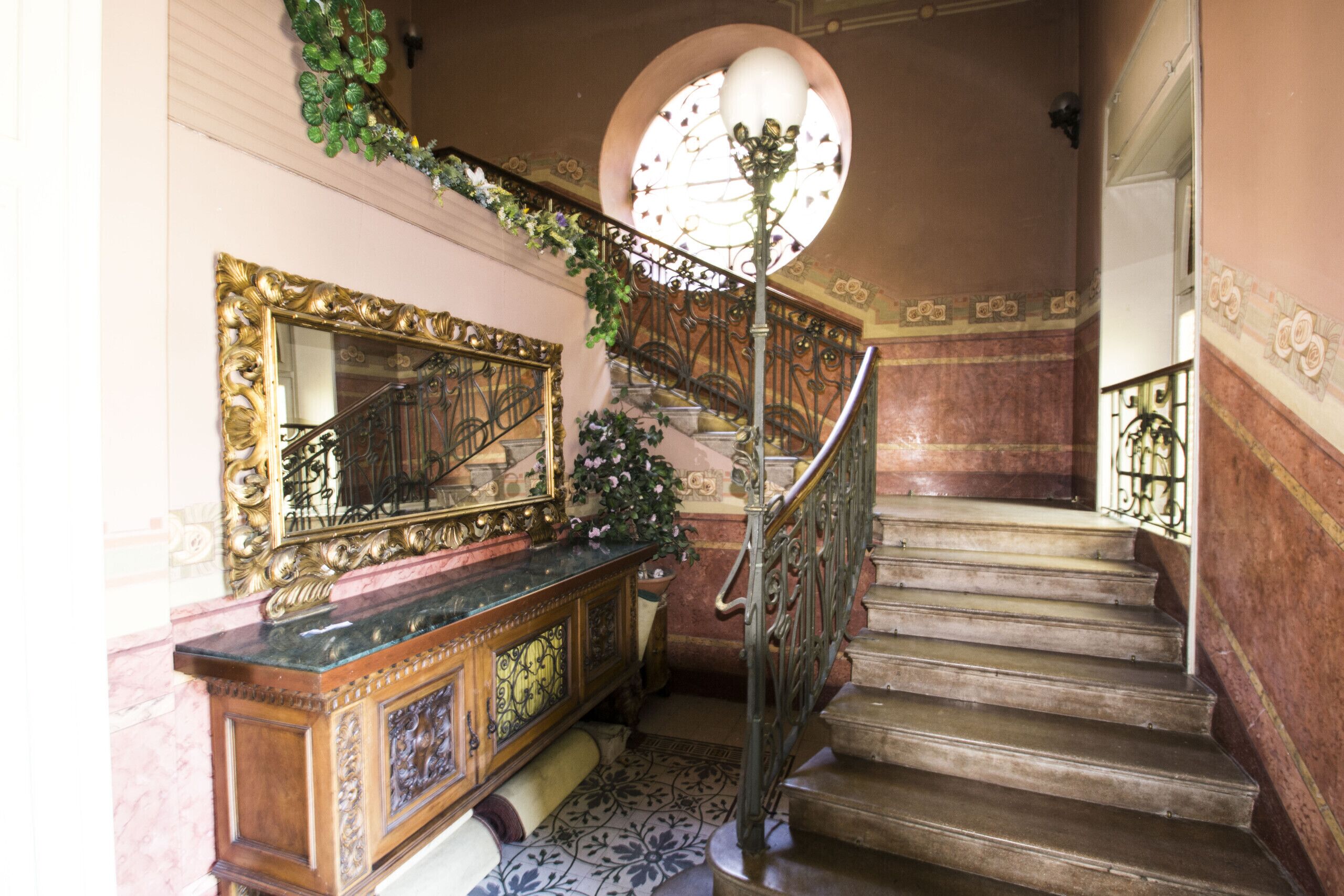 A grand staircase with ornate metal railings, a marble floor, and a large mirror on the wall.