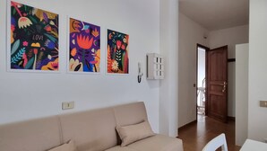A living room with a beige sofa, three colorful wall art pieces, a white wall-mounted phone, and a wooden door leading to another room. - Apartment in La Caletta 5 minutes from the beach (La Caletta)