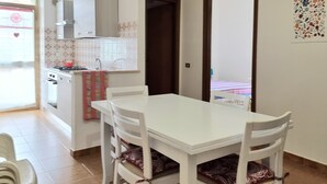 A small dining area with a white table and four chairs, a kitchen in the background, and a door leading to another room. - Apartment in La Caletta 5 minutes from the beach (La Caletta)