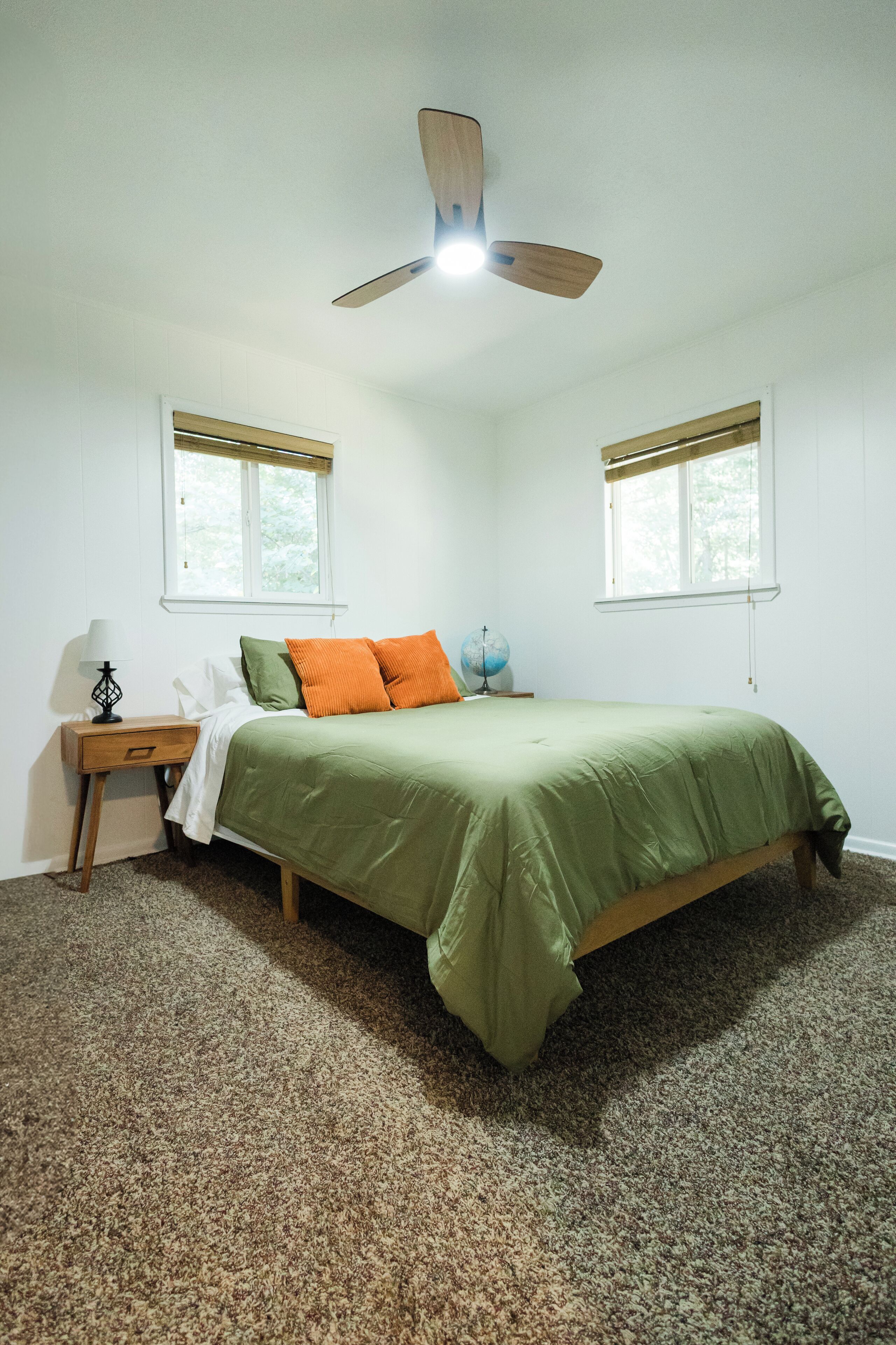 A bedroom with a bed, two windows with blinds, a nightstand with a lamp, and a ceiling fan.