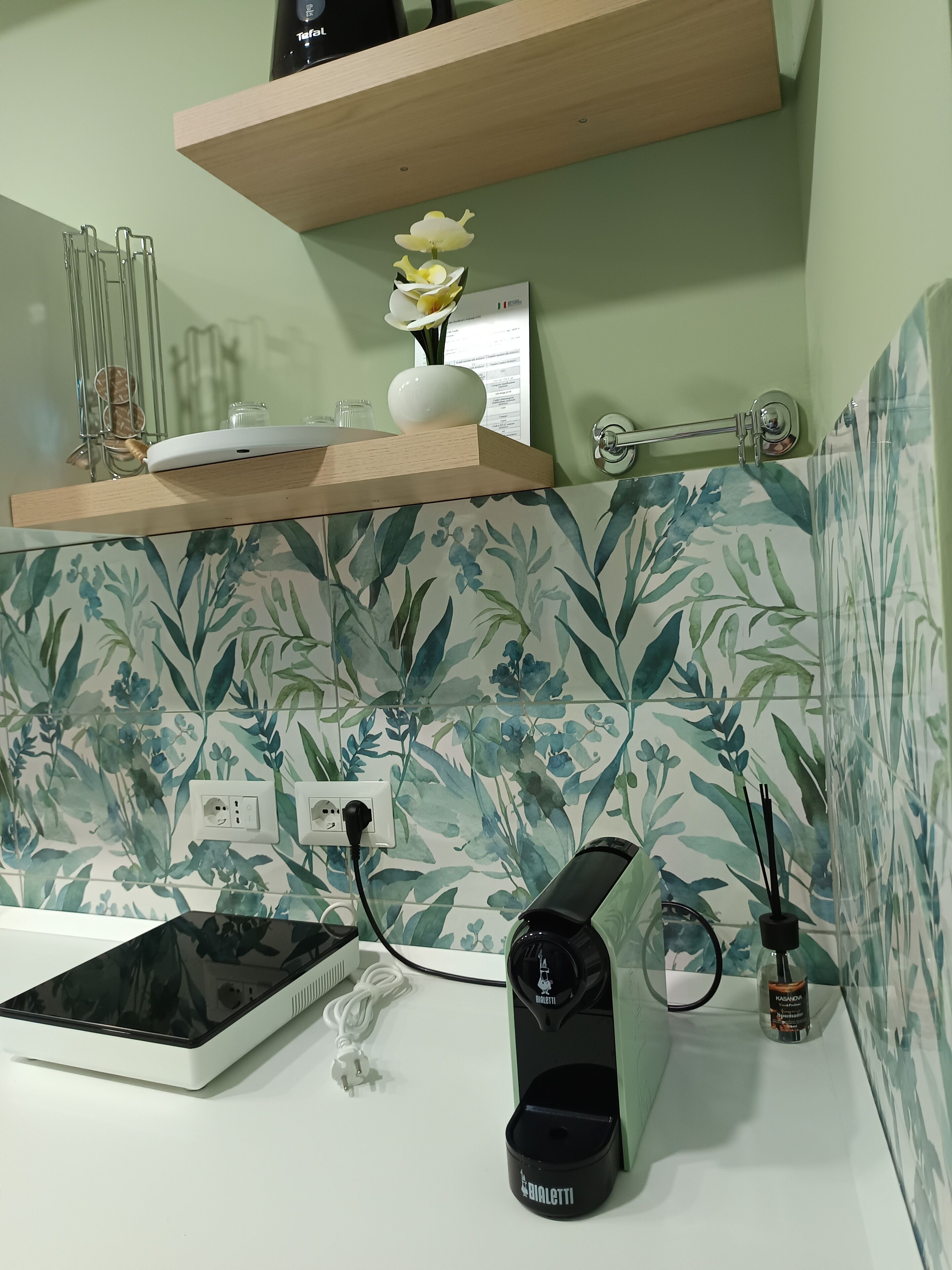 A bathroom with a floral tile backsplash, a white countertop, a black hair dryer, and a small white vase with flowers on a wooden shelf.