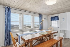 Een eetgedeelte met een houten tafel, bankjes en uitzicht op het strand door grote ramen.