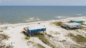 Een pand aan het strand, met een huis op palen, een kleinere constructie en een zandstrand met weinig begroeiing.