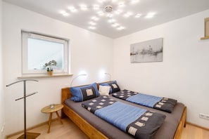 Un dormitorio con dos camas, una ventana con persianas, una mesita de noche, una lámpara de pie y un cuadro en la pared.