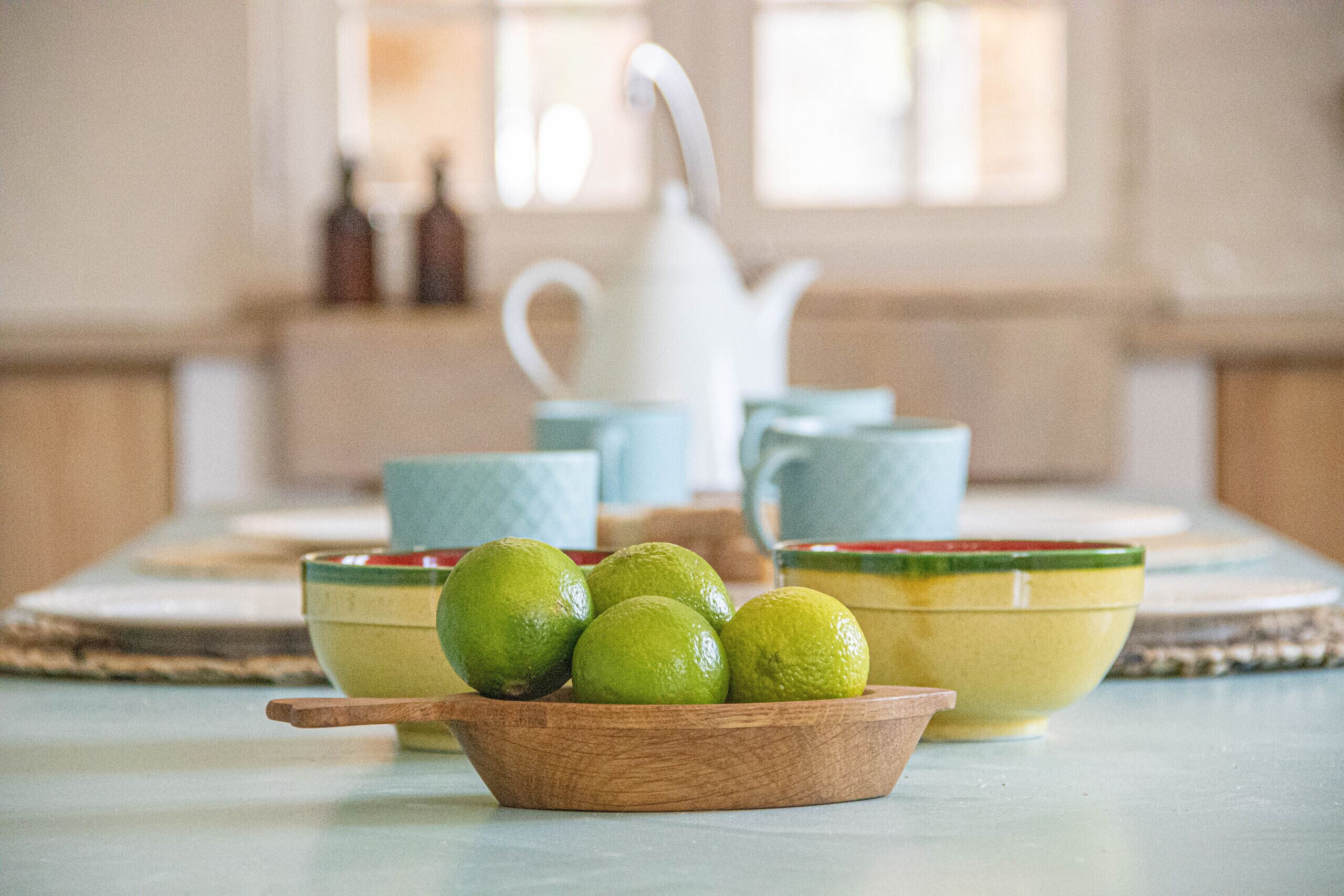 A kitchen with a wooden bowl of limes, a white teapot, and blue cups on a table.