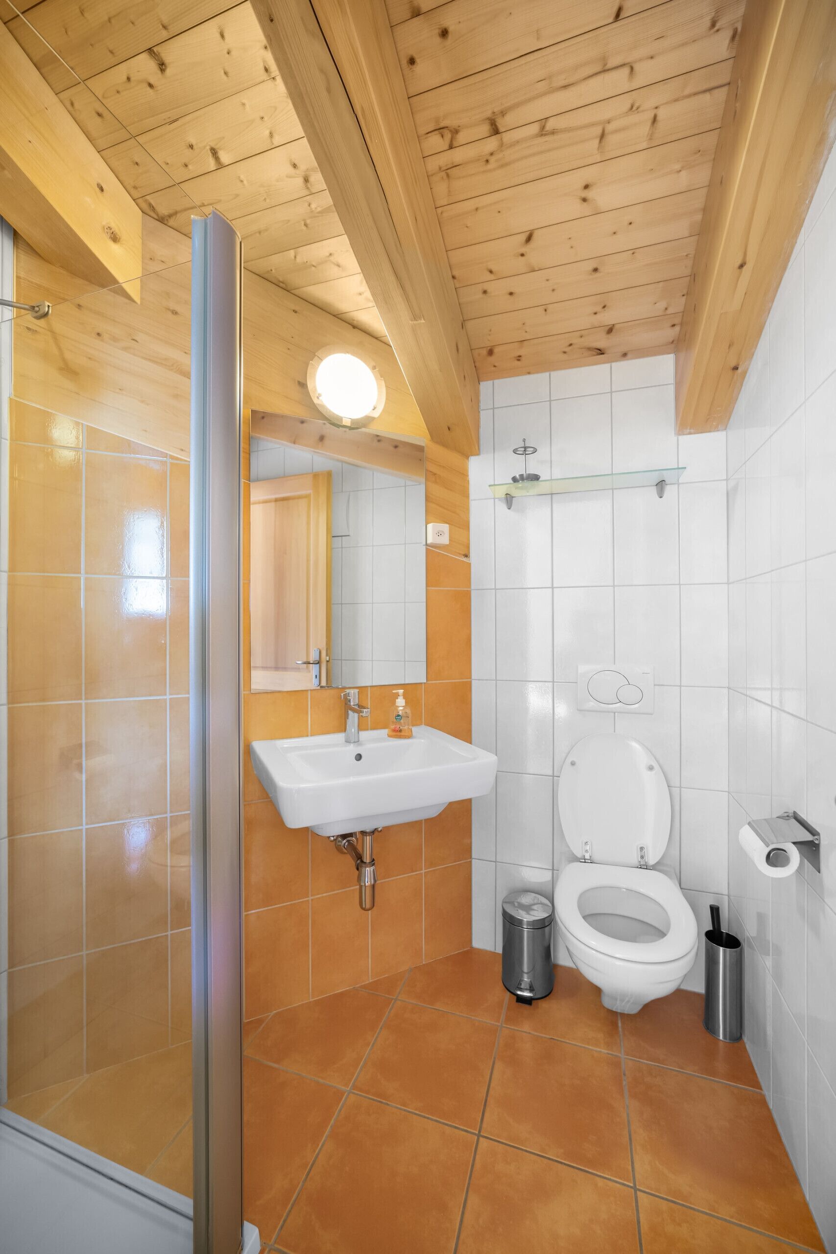 Een badkamer met een houten plafond, een douchecabine met glazen wanden, een wit wastafelmeubel, een toilet en een prullenbak.