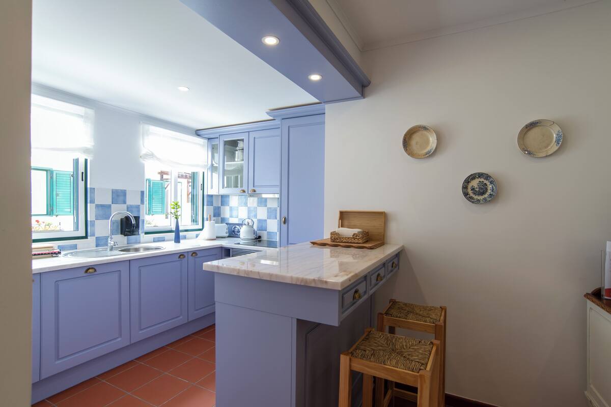 Una cocina con armarios azules, encimera de mármol y una pequeña isla. Se observan dos platos colgados en la pared y una silla de mimbre.
