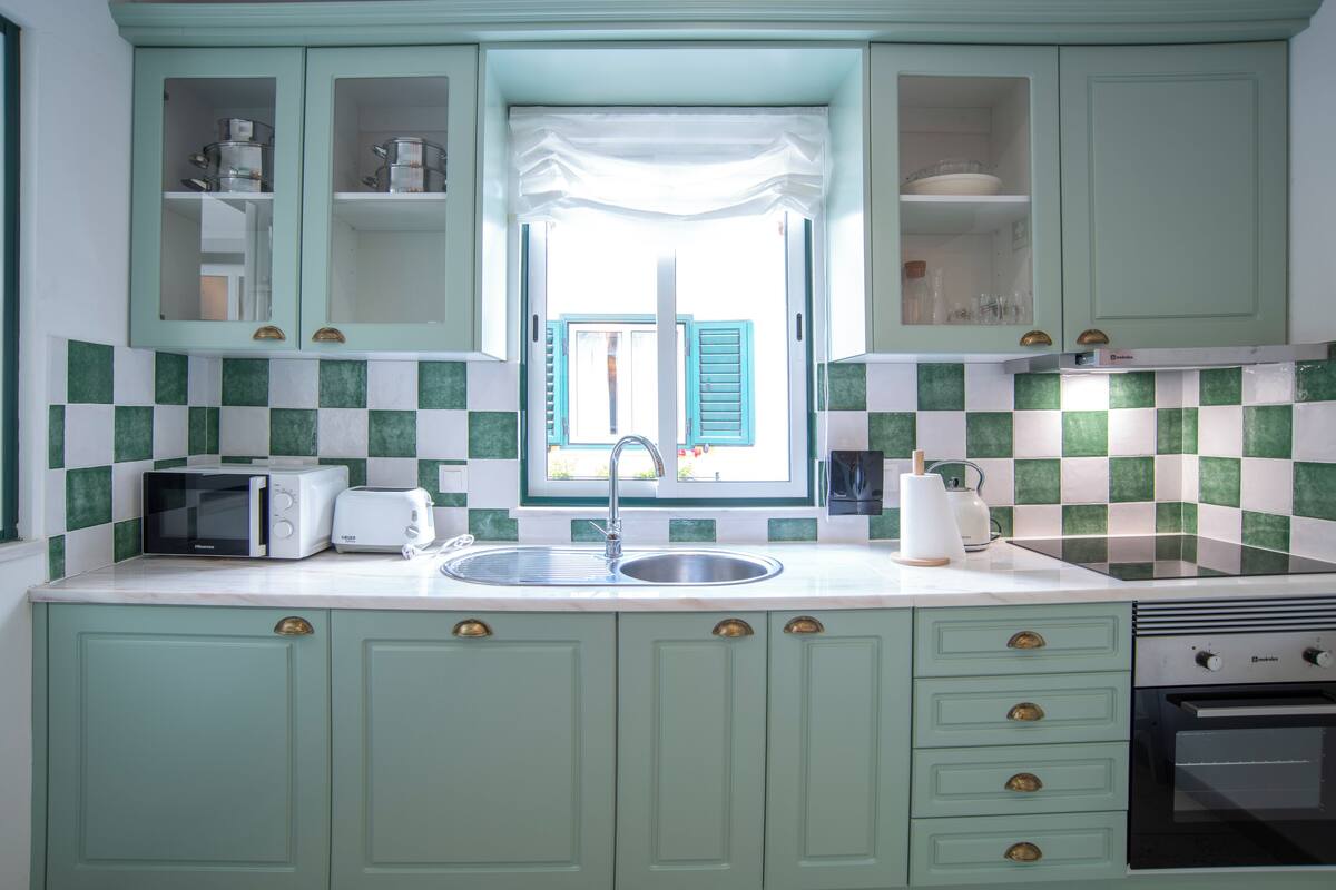 Una cocina con gabinetes verdes, encimera blanca, pileta, tostadora, tetera y estufa.