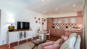 A living room with a TV, white furniture, a glass coffee table, and a kitchen with pink cabinets. - Aposento Real Villas (Montemor-o-Novo)
