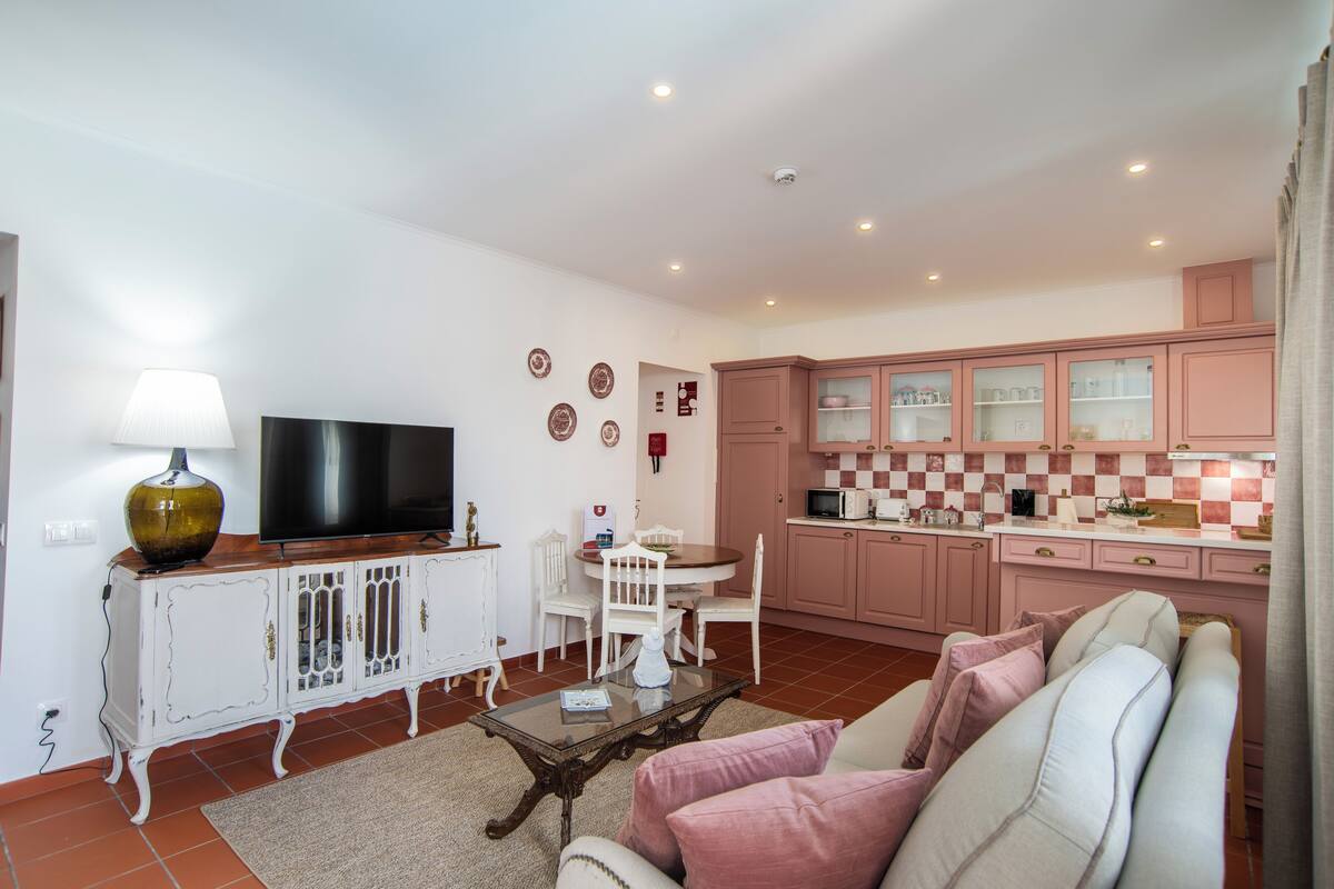 Una sala de estar con televisor, muebles blancos, una mesa de centro de vidrio, y una cocina con gabinetes rosados.