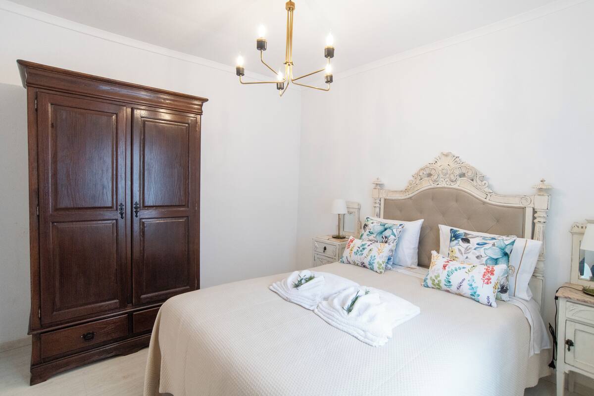 Un dormitorio con un armario de madera grande, una cama con almohadas florales y una araña.