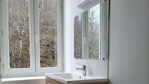 A bathroom with a wooden vanity, a white sink, a mirror, and a window with a view of trees. - Cosy apartment Ideal for hiking and nature (Laruns)