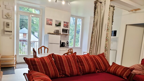 A cozy living room with a red sofa, wooden chairs, a small dining table, and a kitchen area with a refrigerator, microwave, and coffee maker. - Cosy apartment Ideal for hiking and nature (Laruns)
