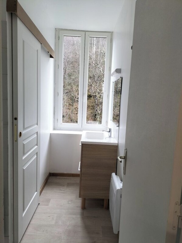 A narrow bathroom with a window, a white sink, and a wooden cabinet. - Cosy apartment Ideal for hiking and nature (Laruns)
