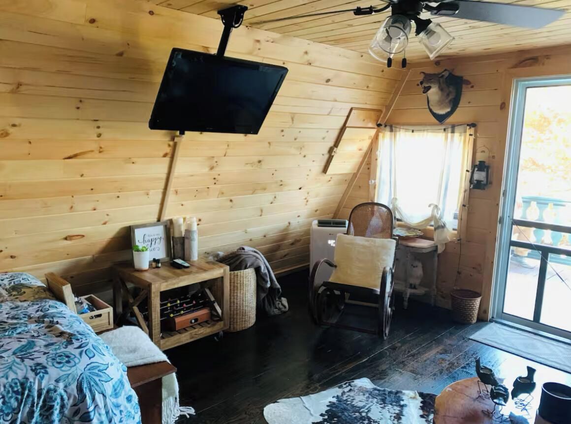 A cozy cabin interior with a wooden ceiling, a mounted TV, a wheelchair, and a window with a view of the outdoors.