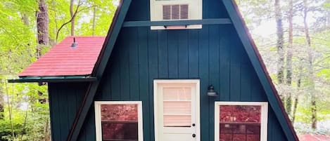 Une petite cabane triangulaire, de couleur vert foncé, avec un toit rouge, une porte blanche et deux fenêtres de chaque côté.