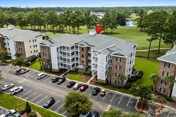 A multi-story hotel with a parking lot and a golf course in the background. - Golf & Sunshine - Spacious Condo Overlooking the Green (Myrtle Beach)