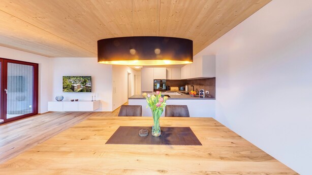 A modern kitchen with a central island, a dining area with a wooden table, and a large window with a view of a building. - Urban-Apartmenthaus zwischen Berg und See (Dornbirn)