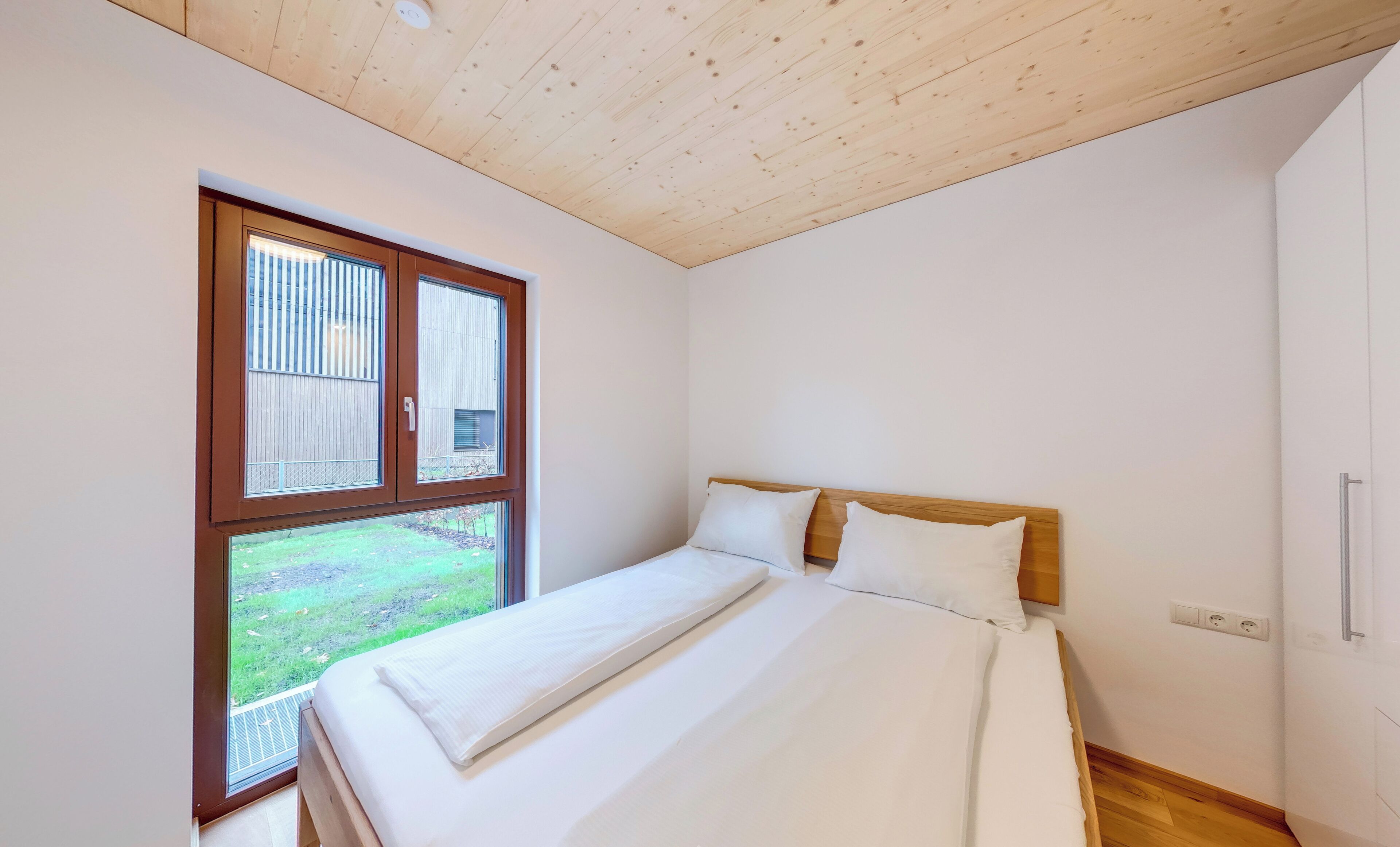 Una camera da letto con soffitto in legno, un'ampia finestra, un letto con biancheria bianca e pavimento in legno.
