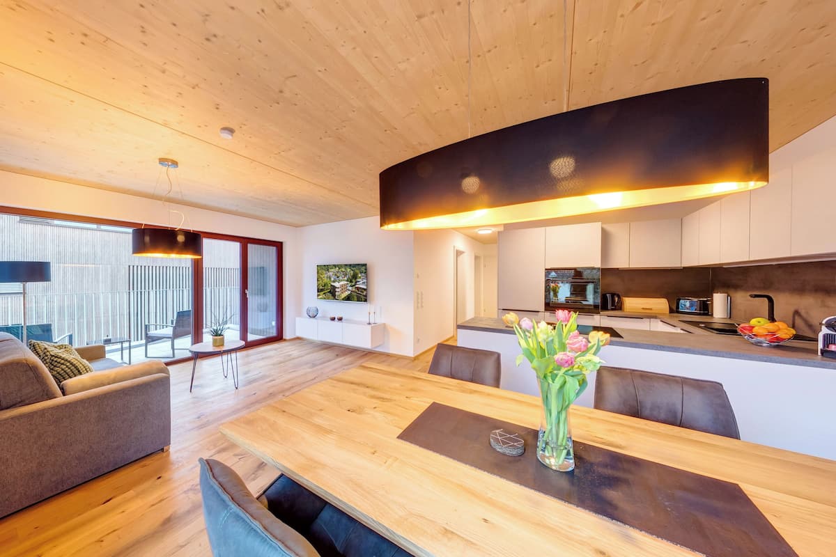 A modern living space with a wooden dining table, sofas, and a kitchen area with a TV and fruit bowl.