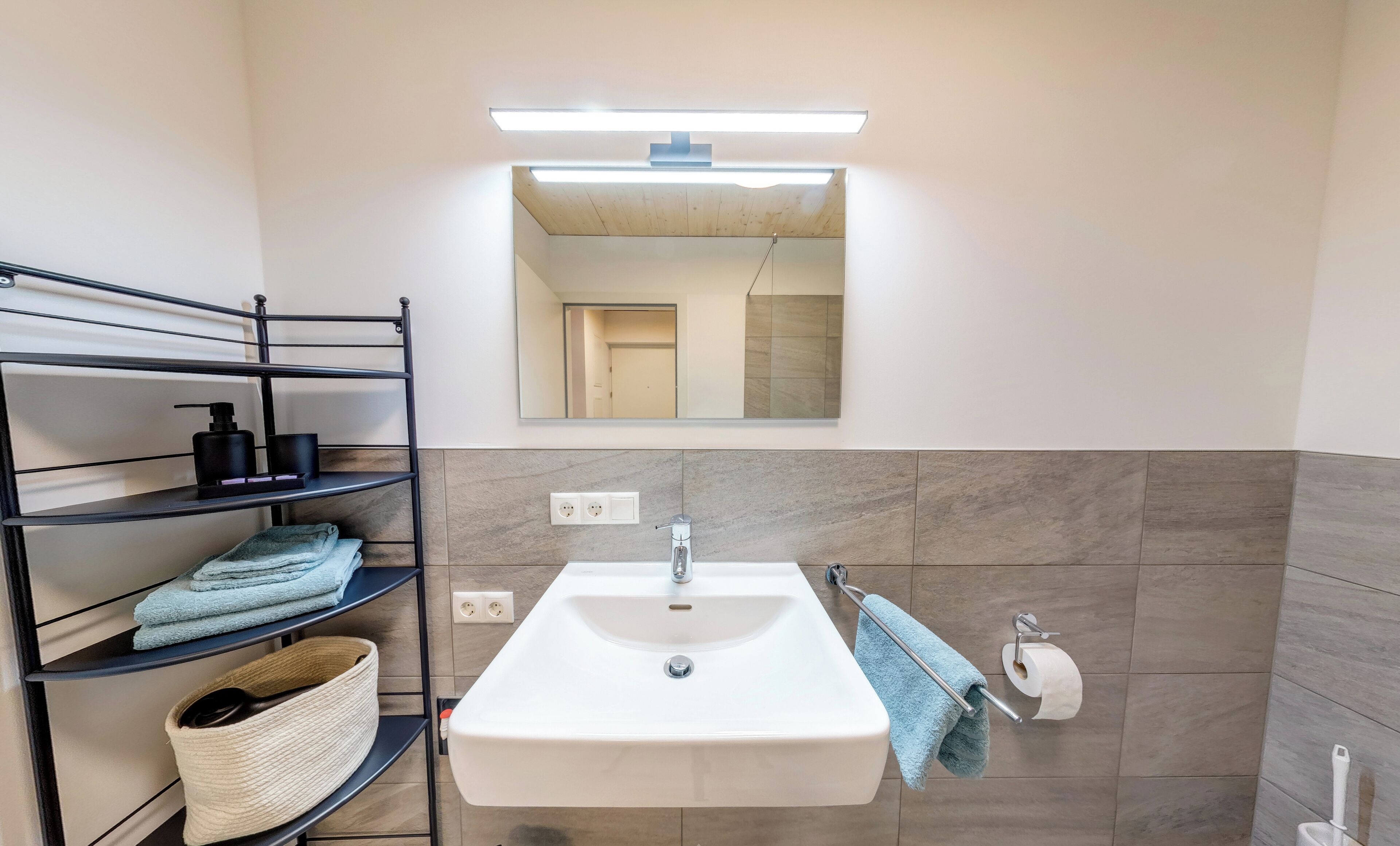 Ein Badezimmer mit einem weißen Waschbecken, einem Spiegel und einem Handtuchhalter. Regale mit Handtüchern und Toilettenartikeln sind vorhanden.