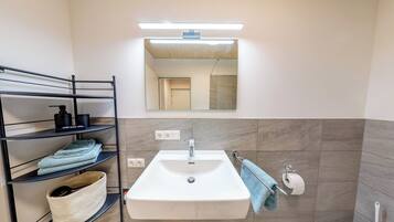 Ein Badezimmer mit einem weißen Waschbecken, einem Spiegel und einem Handtuchhalter. Regale mit Handtüchern und Toilettenartikeln sind vorhanden.