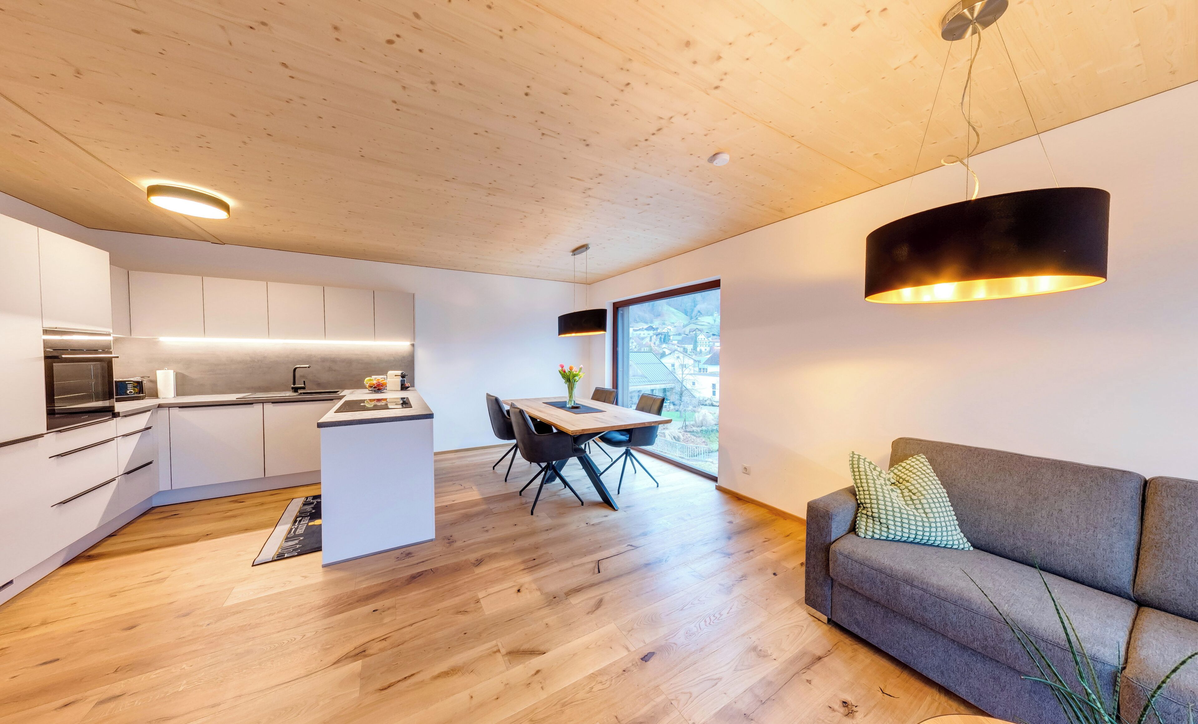 Una cocina moderna con una isla central, una zona de comedor con mesa y sillas, y un sofá gris con una planta.