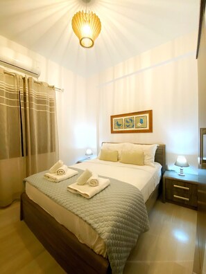 A hotel room with a bed, a bedside table, a lamp, a framed picture on the wall, and a window with curtains.