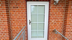 A white-framed door with a window, set in a brick wall with a metal handrail and a light fixture above. - Charming top floor apartment near the Baltic Sea Ideal for families of up to 6 people (Rövershagen)