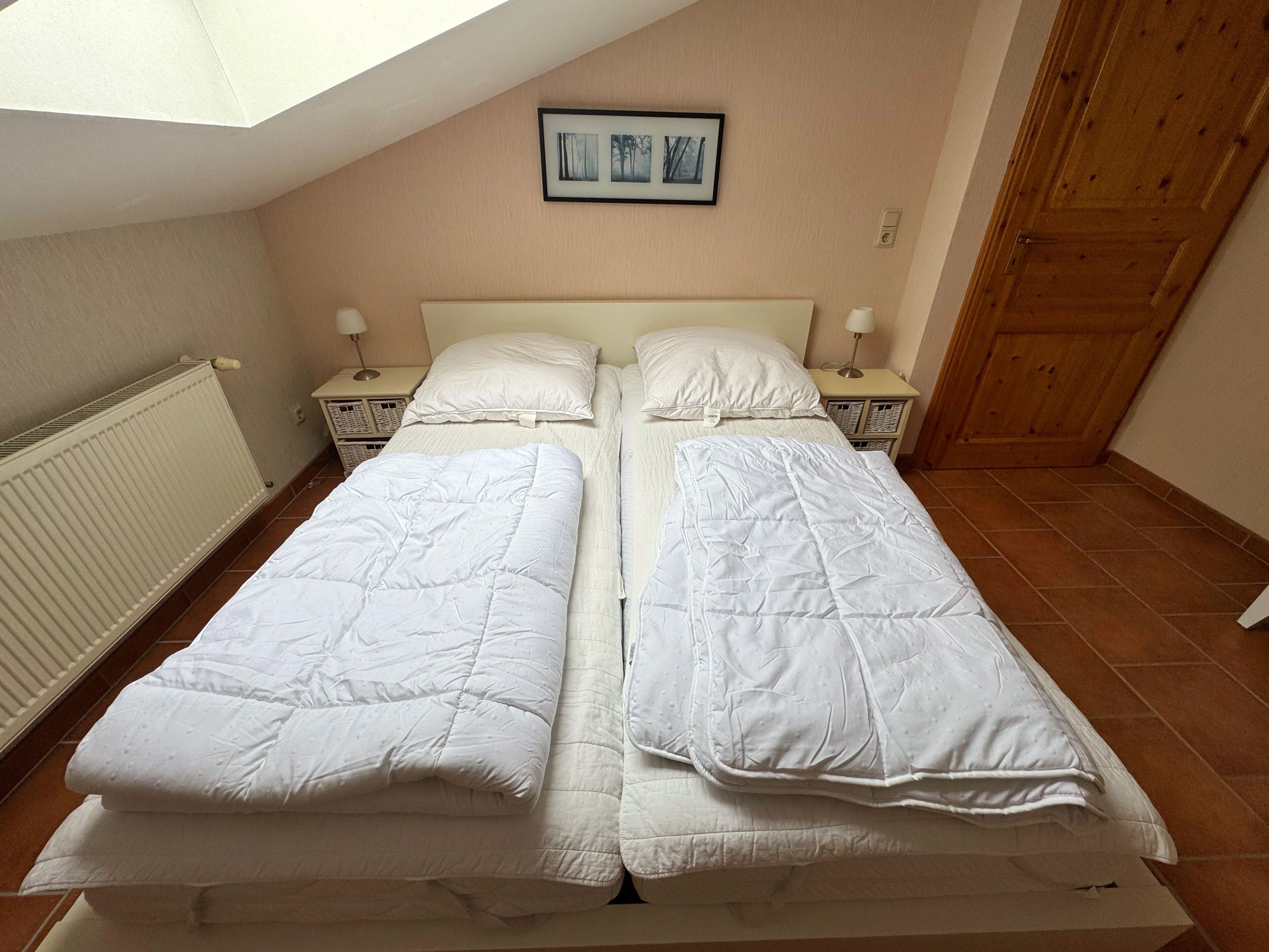 Una cama con dos plazas, ropa de cama blanca, una puerta de madera, un radiador y un cuadro en la pared.