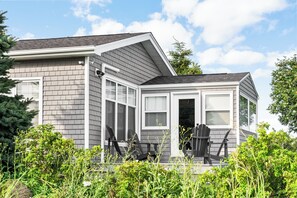 A modern house with a grey exterior, large windows, and a covered porch with two chairs. - Premium Oceanfront Cottage Near Shediac | Modern Comforts & Stunning Views (Grande-Digue)