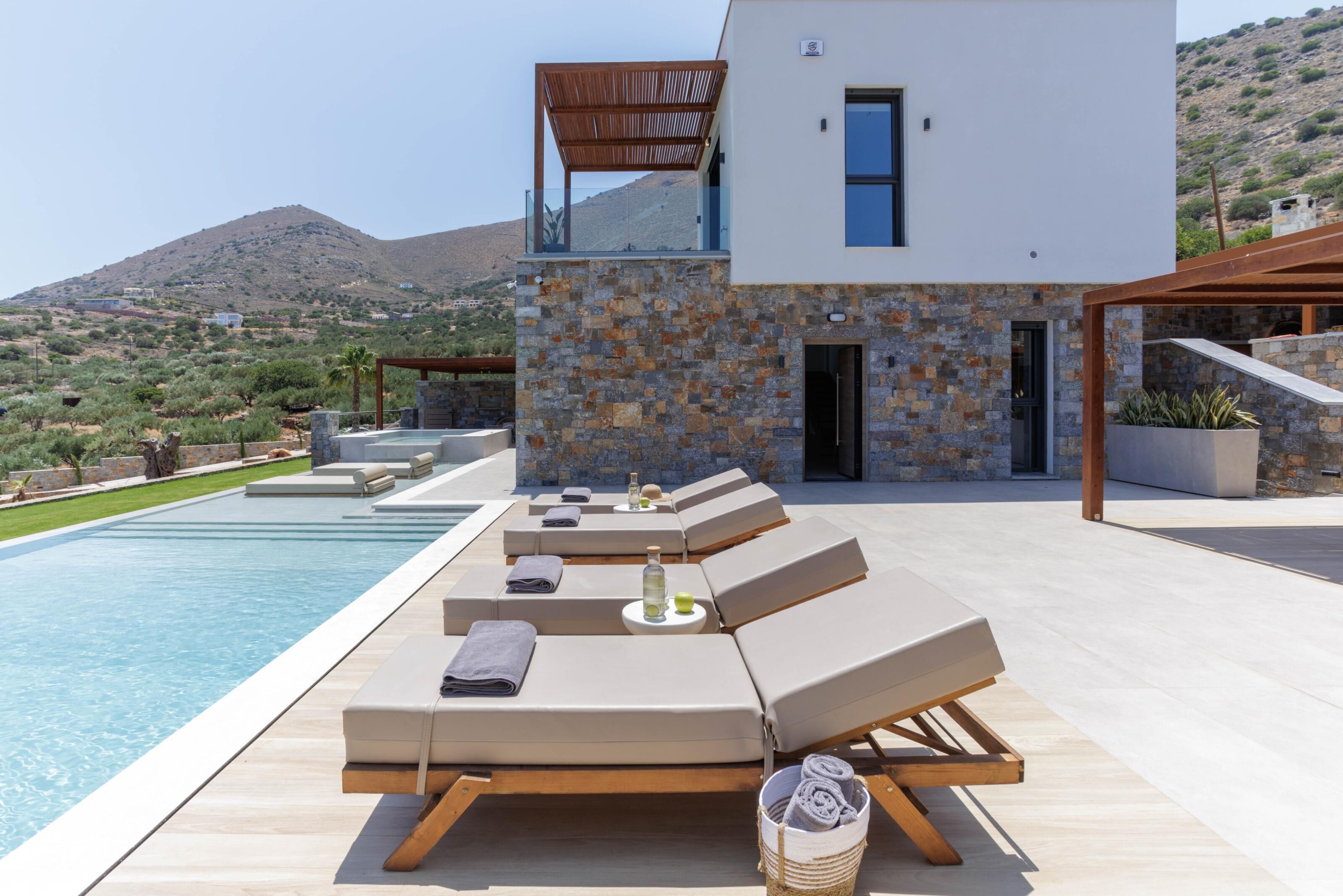 A modern outdoor pool area with lounge chairs, a stone wall, and a view of a mountainous landscape.