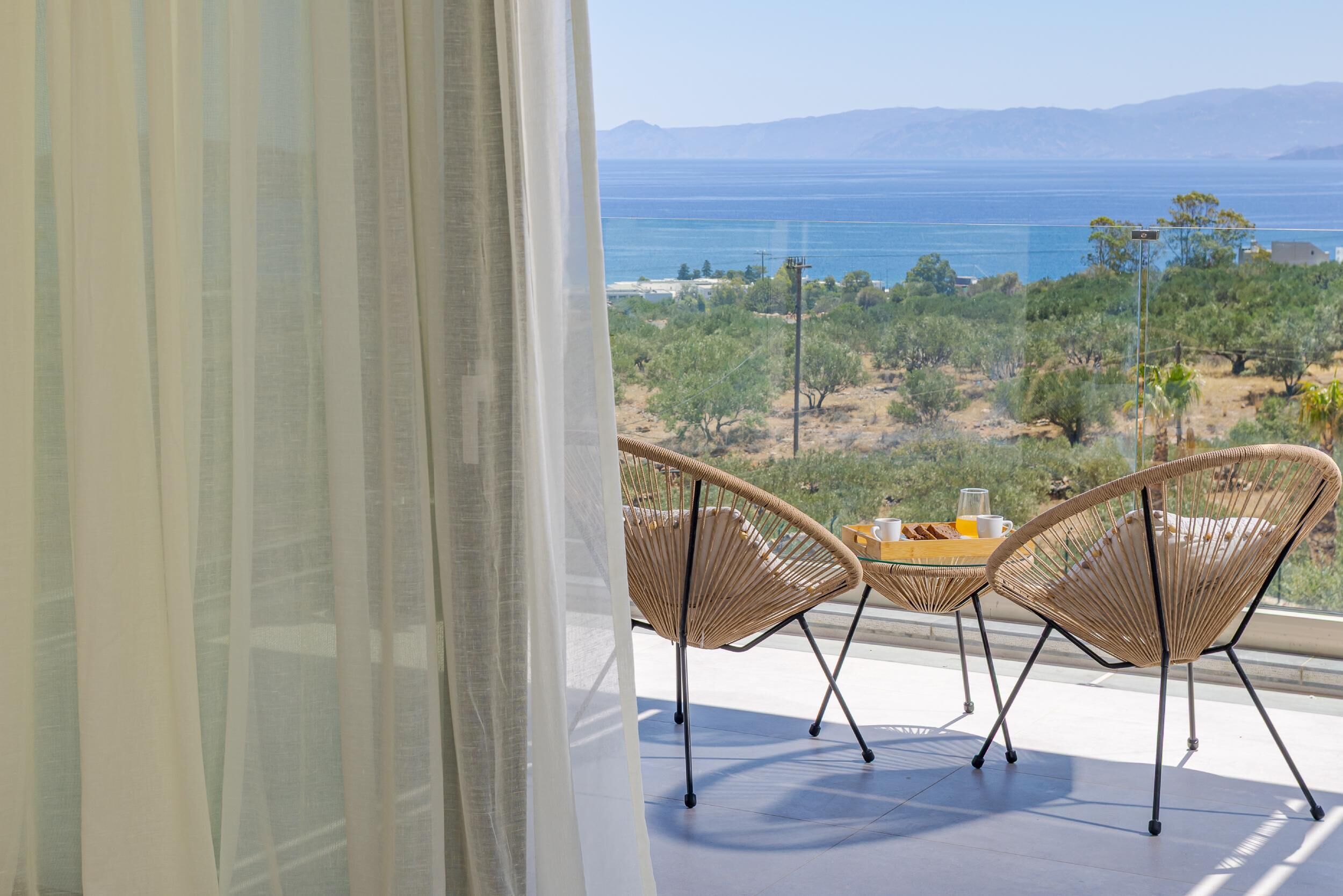 Un balcon agrémenté de chaises et d’une table en osier, offrant une vue sur un paysage côtier avec des arbres et la mer.