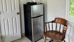 A corner room with a white door, a stainless steel refrigerator, a wooden chair, and a window with a view of greenery. - Glades Motel (Naples)