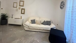 A living room with a white sofa, a side table, a potted plant, and a clock on the wall.