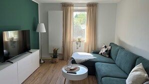En moderne stue med et fladskærms-tv, en grøn sofa, et hvidt sofabord og et vindue med gardiner.