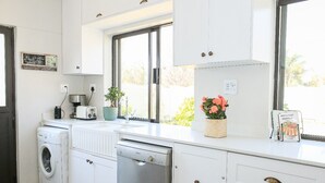 A modern kitchen with white cabinetry, a built-in dishwasher, a washing machine, and a window with a view of greenery. - Kirrin House (Benoni)
