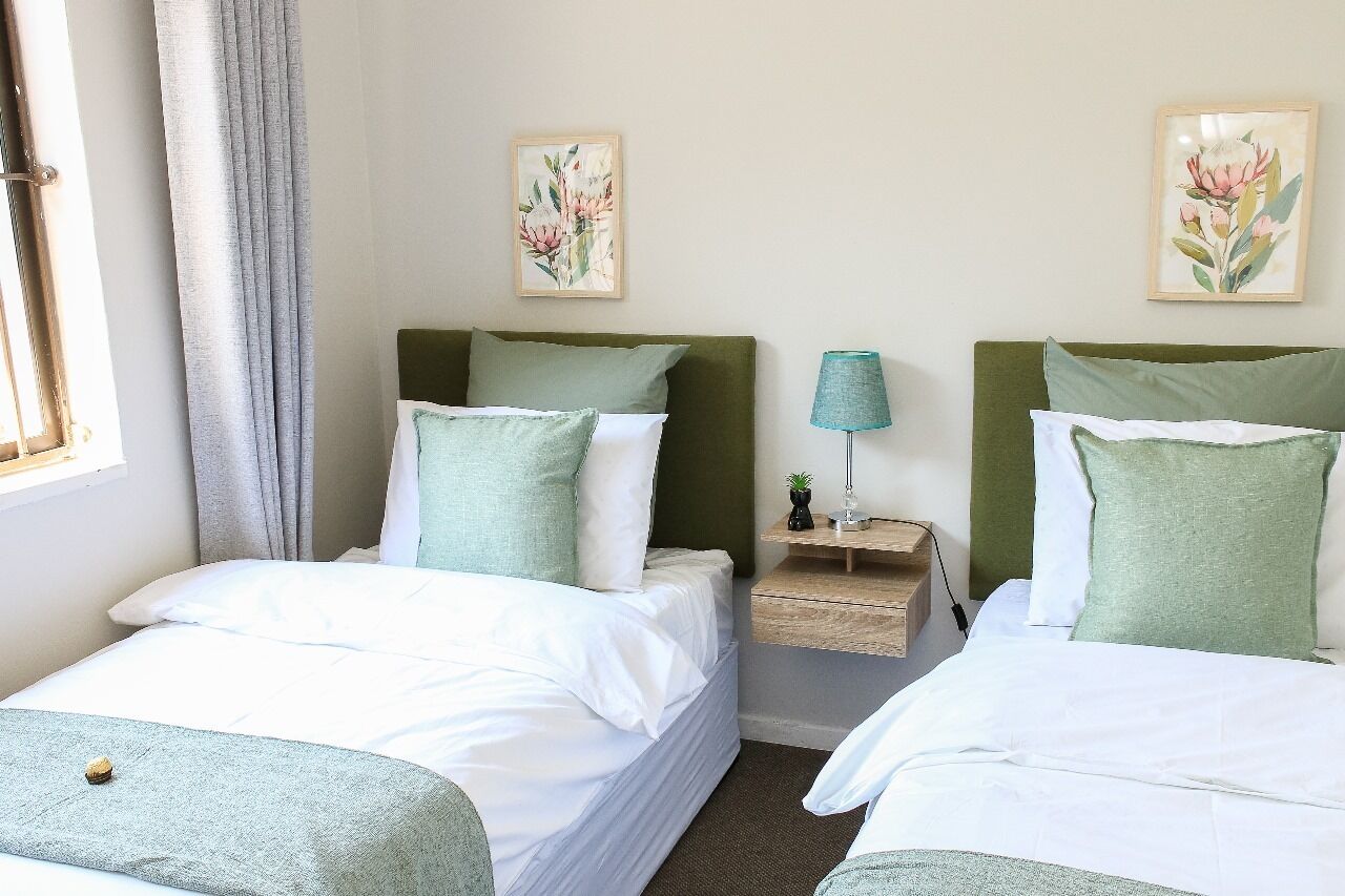 Une chambre d’hôtel avec deux lits, chacun agrémenté de draps blancs et d’oreillers verts. On y trouve une table de chevet avec une lampe et une petite plante. La chambre est dotée d’une fenêtre avec des rideaux, et deux tableaux représentant des motifs floraux encadrés ornent le mur.
